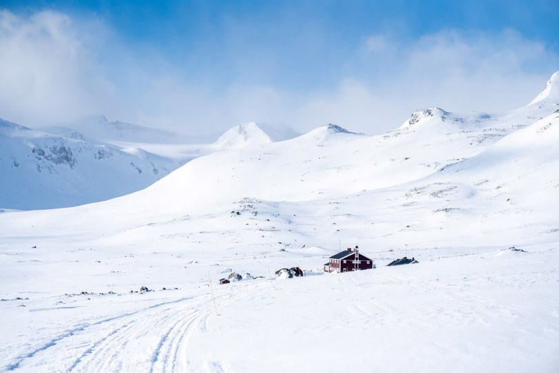 snødekt fjellandskap hvor man ser noen hus et stykke unna. Det er blå himmel