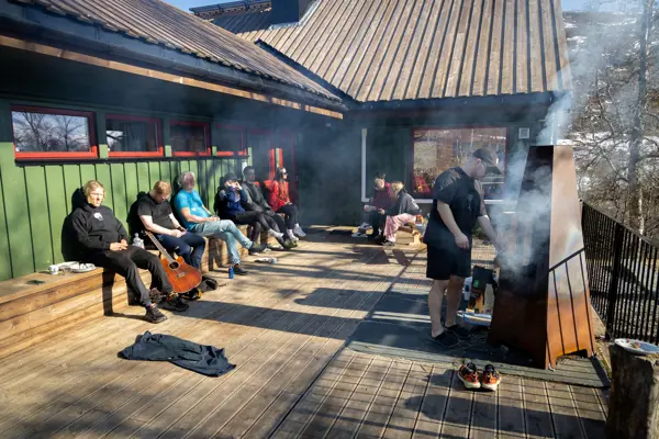 Folk slapper av på terrasse utenfor hytte i solen mens en mann griller.