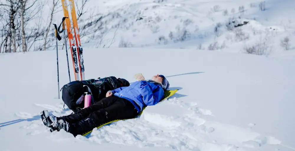 En person som ligger i snøen og soler seg. Det står et par med ski ved siden av