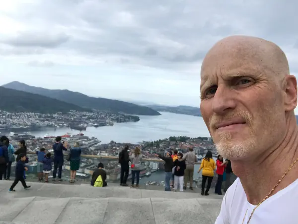 Man i hvis t-skjorte ser inn i kamera på toppen av Fløyen med Bergen bak.
