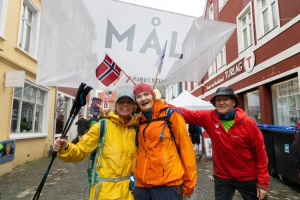 To glade turgåere i mål med en frivillig som holder opp norsk flagg bak dem.