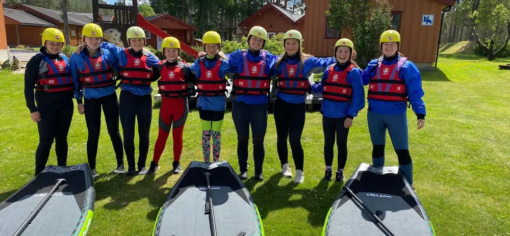 Ungdommer står på rekke foran paddleboards, de har på seg hjelm og redningsvest. 