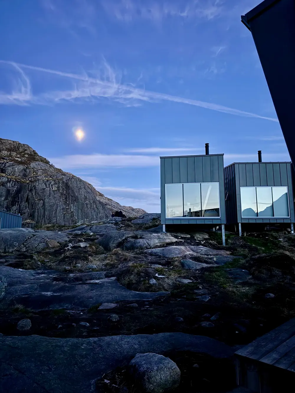 En grå hytte med store vinduer som ligger på fjellet. Måneskinn og kveldslys.