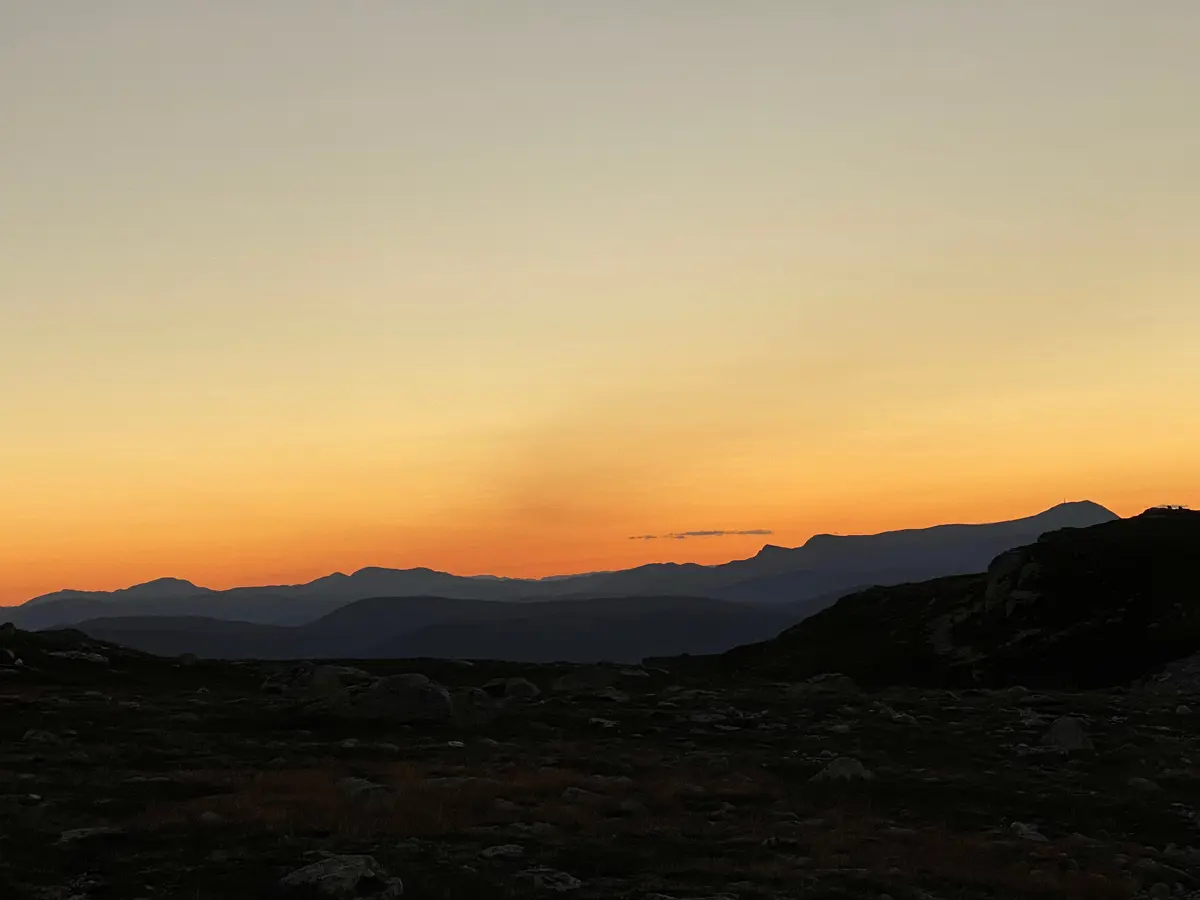Utsikt over fjelllandskap i solnedgang. Himmelen er oransje og man ser kun konturene av fjell.