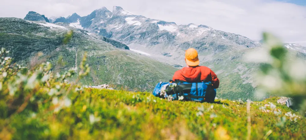 En mann i rød jakke og gul caps som sitter i lyngen og ser på fjell
