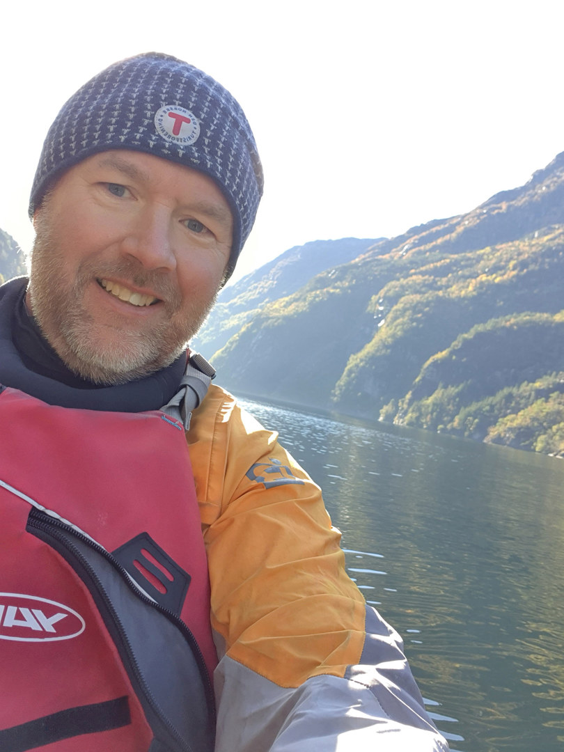Mann med skjegg og lue og redningsvest smiler.