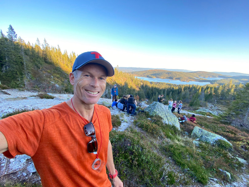 Mann som tar en smilende selfie. Har har på seg en caps med T på forsiden. Han står utendørs i fjell- og skogområde, og det er sol