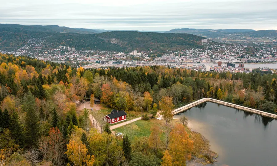liten rød hytte som ligger i skogen ved et vann. i bakgrunnen ser man utsikt til en større by.