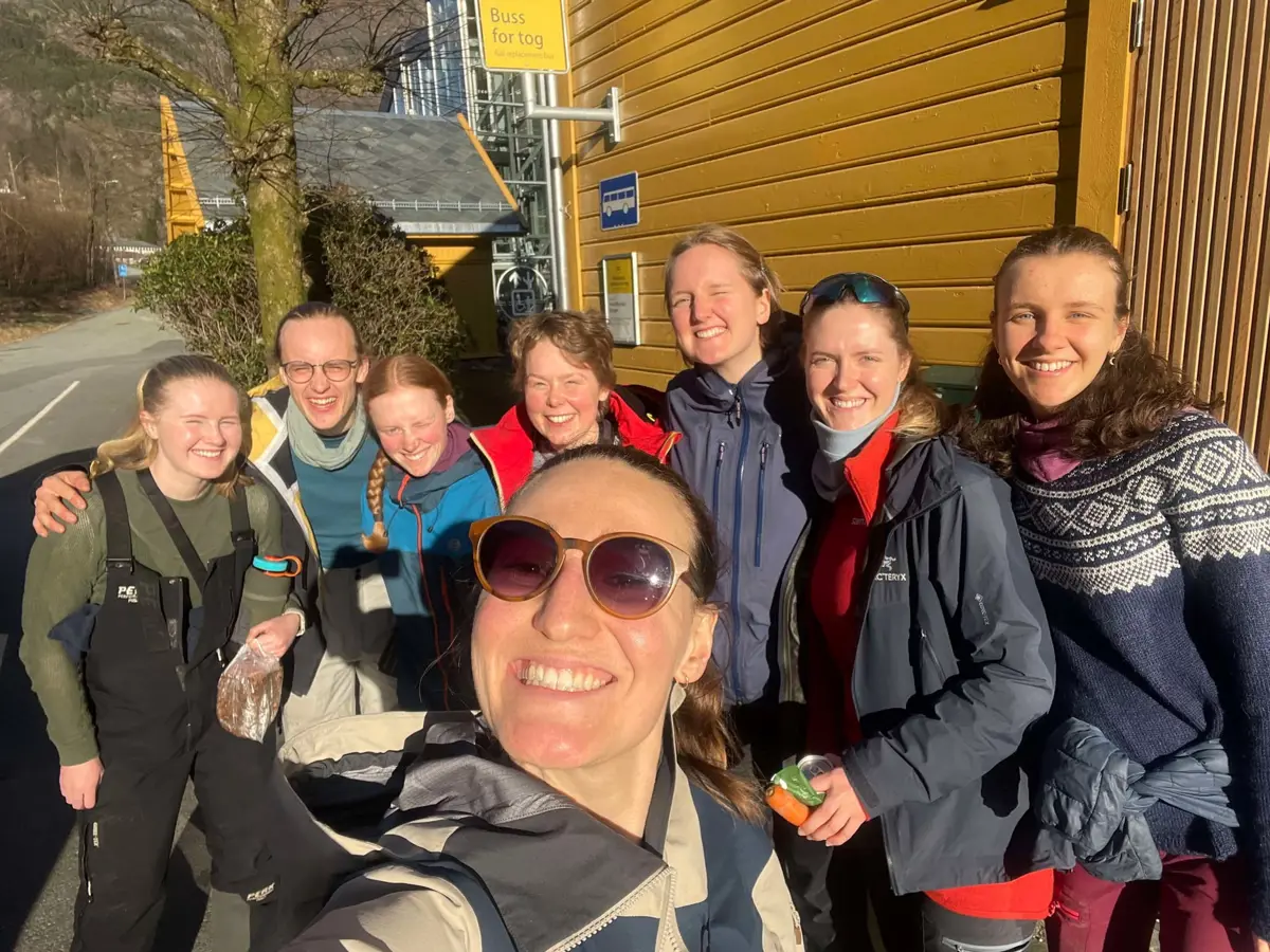 Kvinne med solbriller står og smiler foran gruppe med turkledde mennesker.