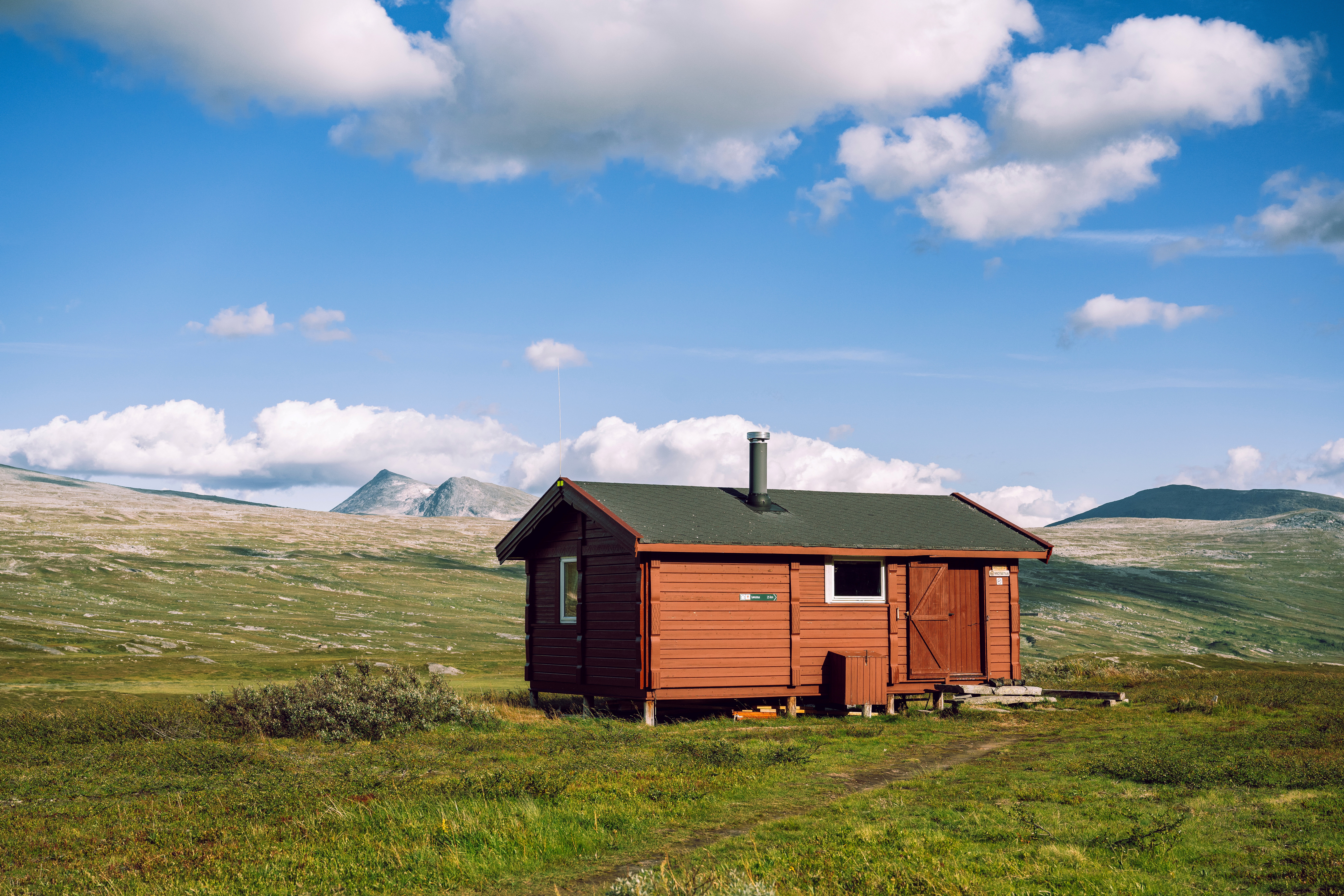 liten r&oslash;d hytte som ligger &aring;pent til i et fjellomr&aring;de. det er bl&aring; himmel og hvite skyer