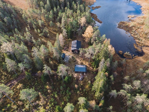 Bilde tatt av drone. Vi ser hytte som ligger ved et vann i skogen.
