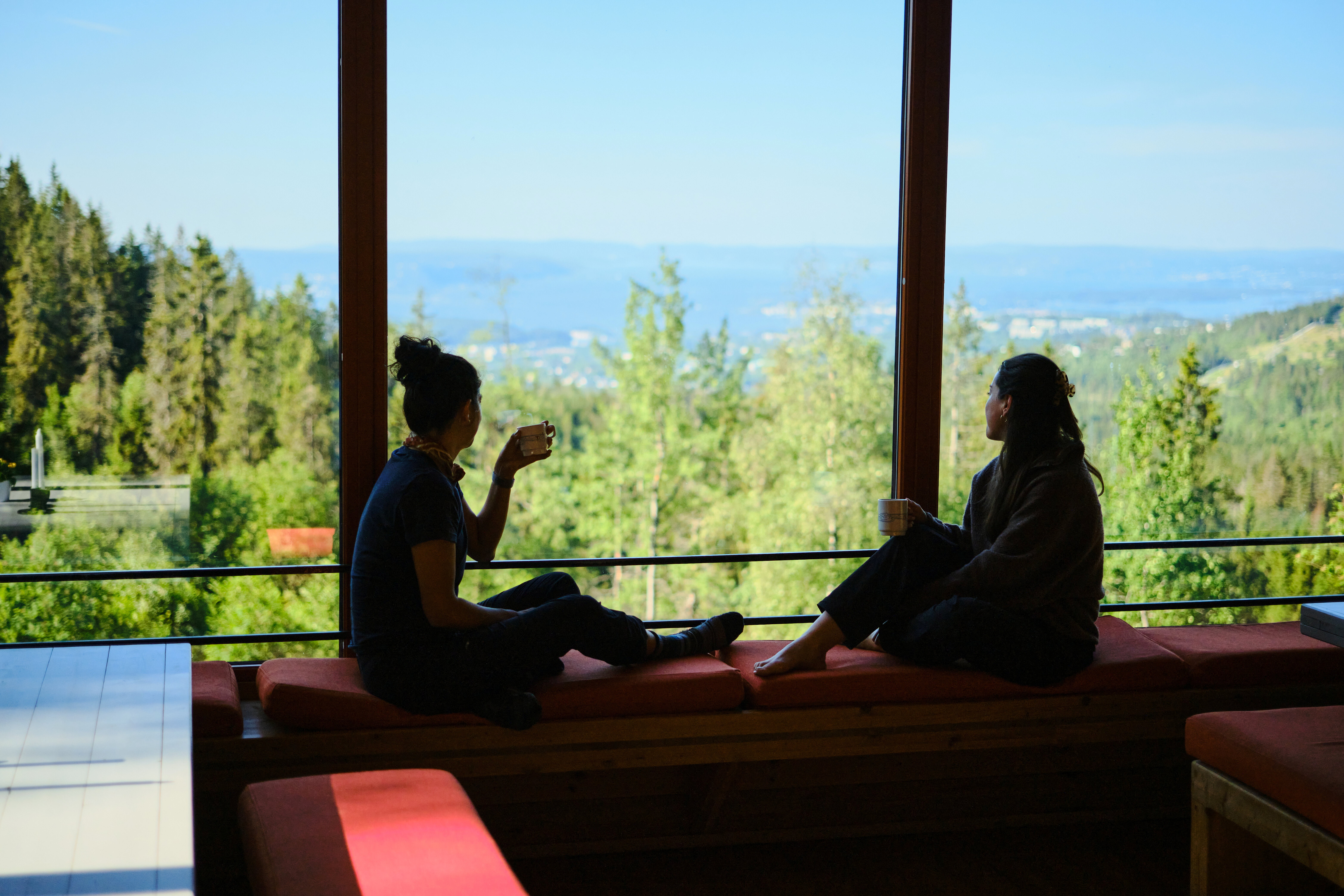 To personer som sitter i et stor vindu og ser utover en grøn skog og oslofjorden