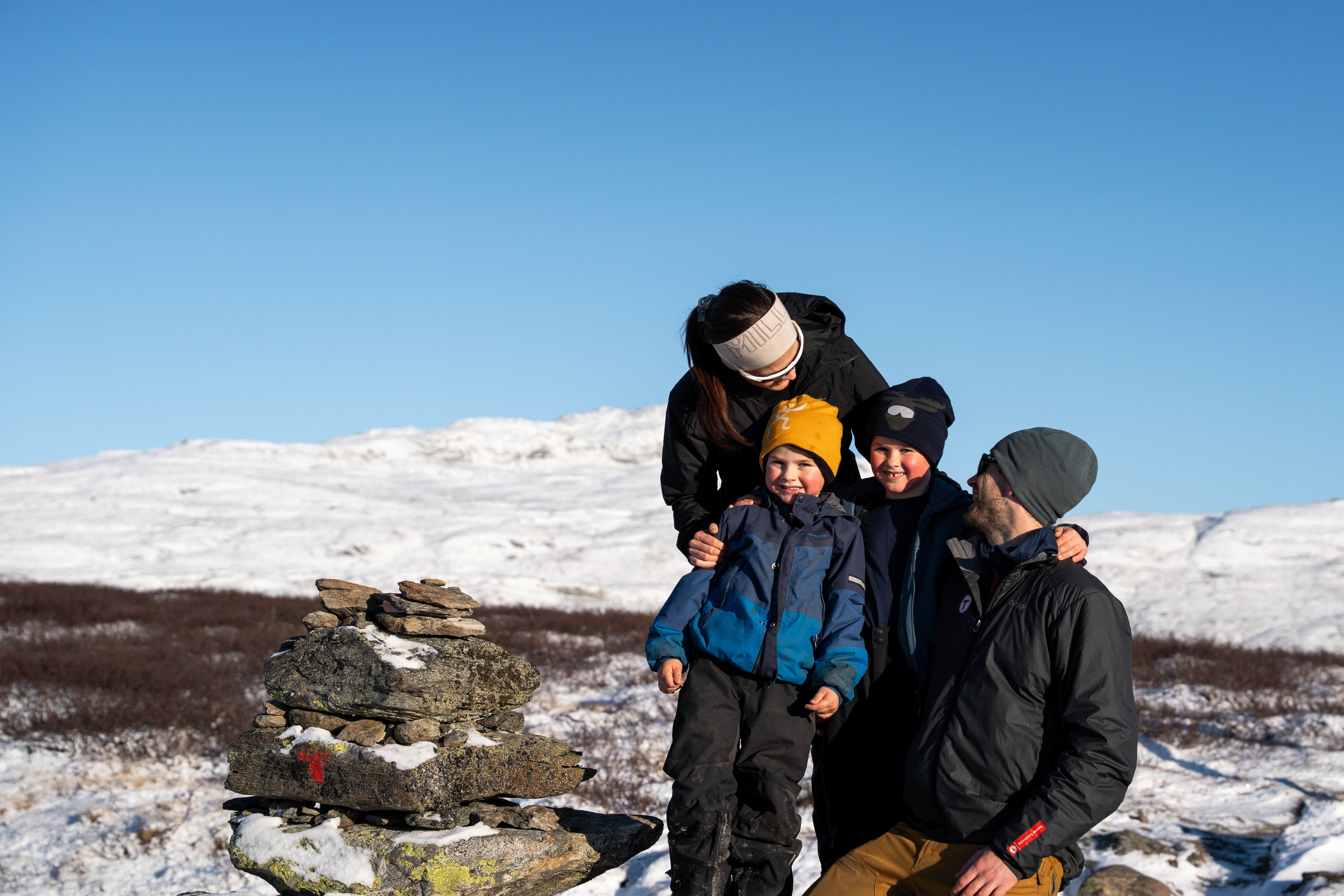 En familie med en mor, far og to gutter som holder rundt hverandre og smiler. Ved siden av dem er det en varde med r&oslash;d T