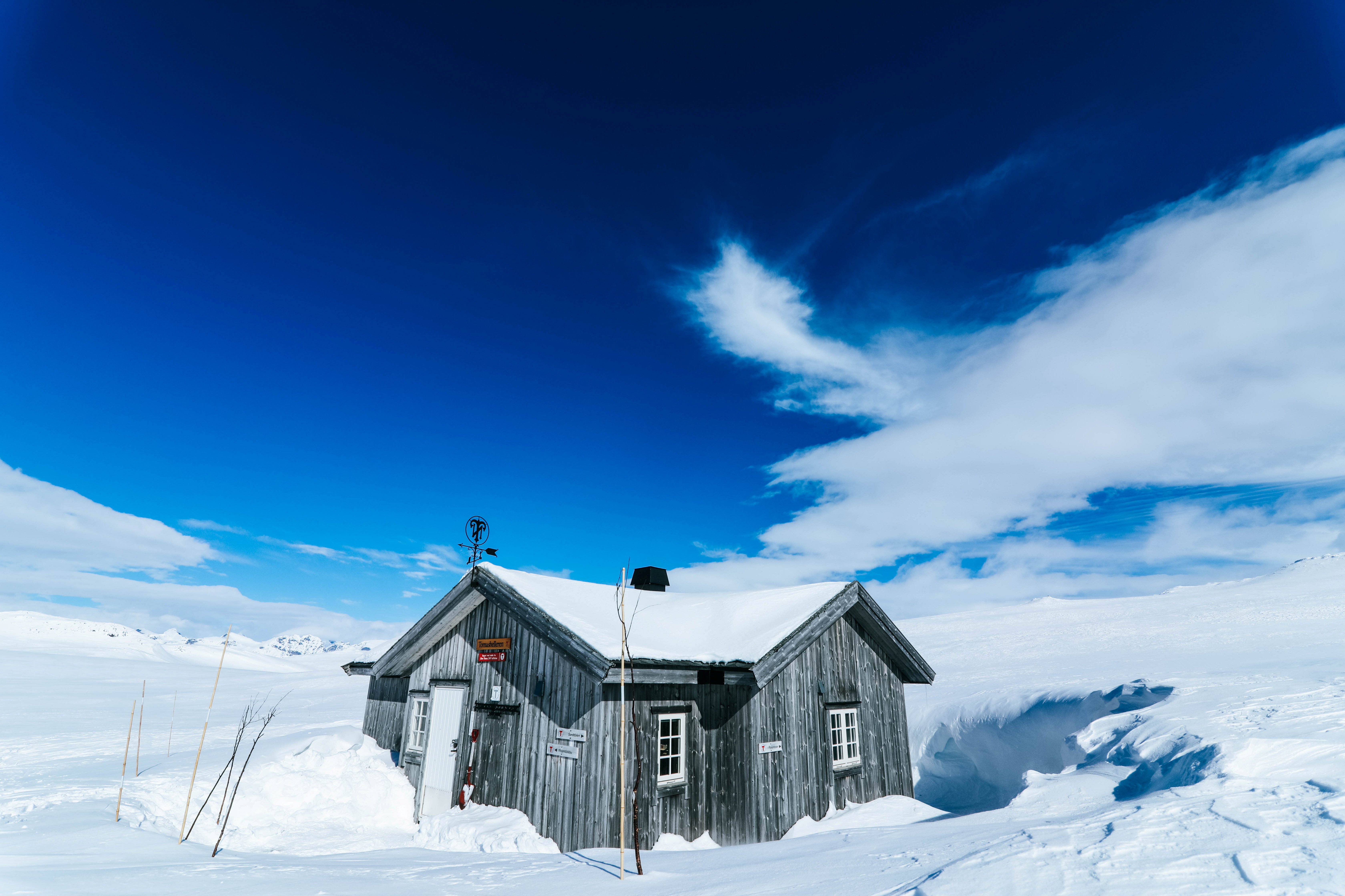 en liten hytte som ligger i et sn&oslash;dekket fjellandskap. Det er bl&aring; himmel og noen hvite skyer