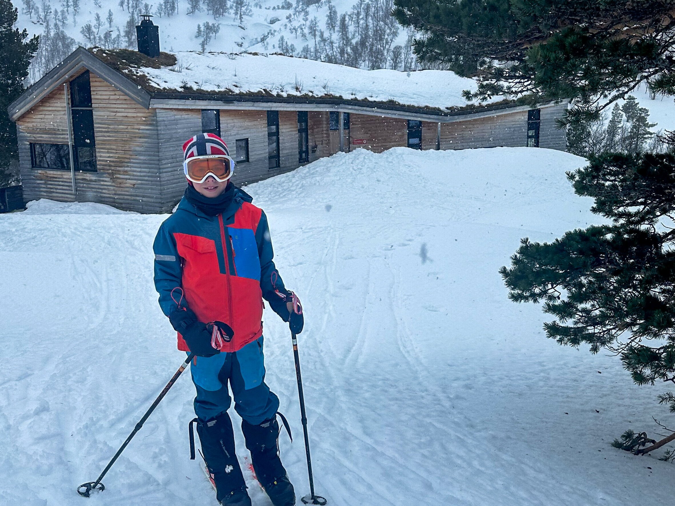 Bilde av en gutt som st&aring;r p&aring; ski utenfor en trefarget hytte.