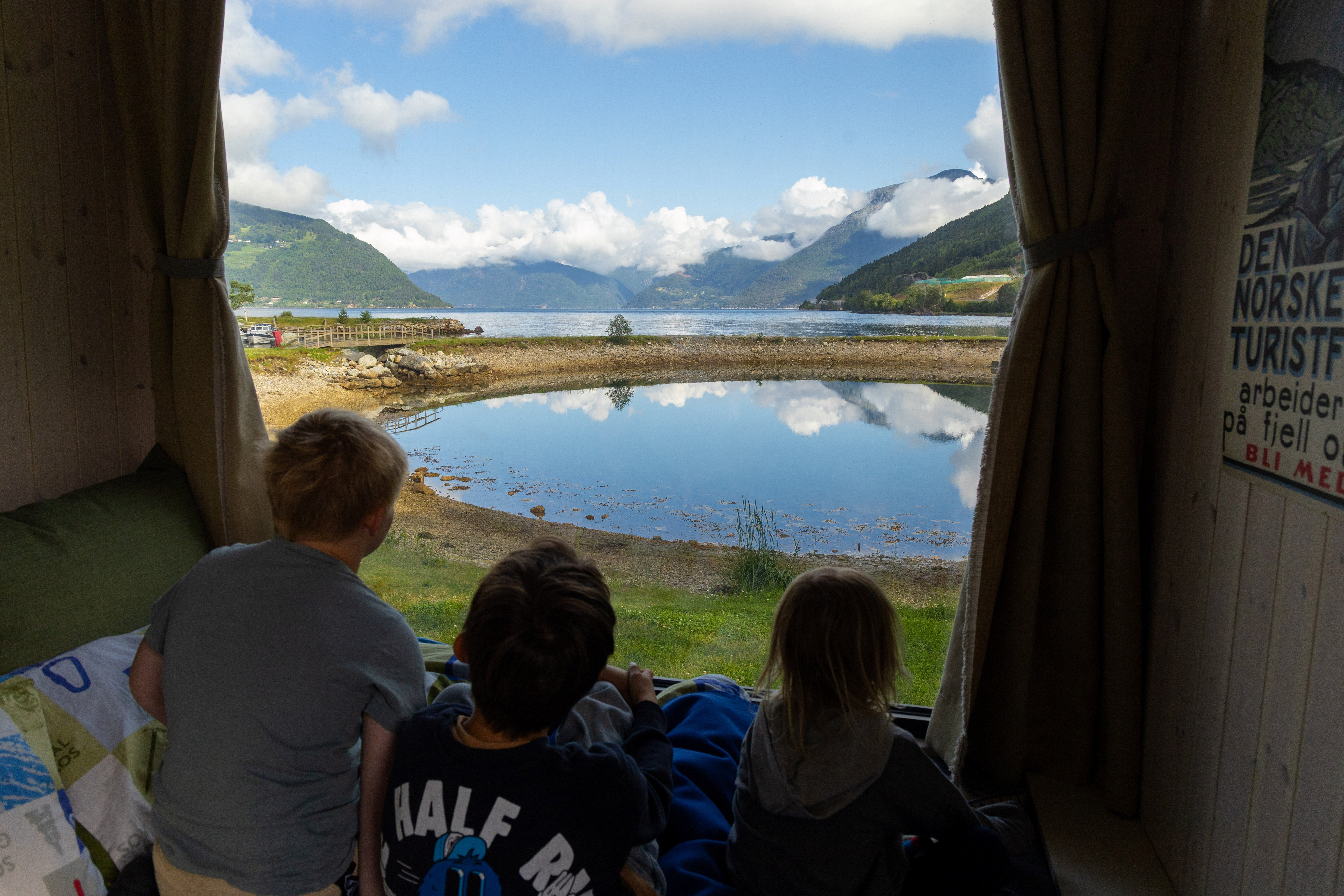 tre barn ser ut gjennom stort vindu mot fjord og fjell. Hytta p&aring; hjul.