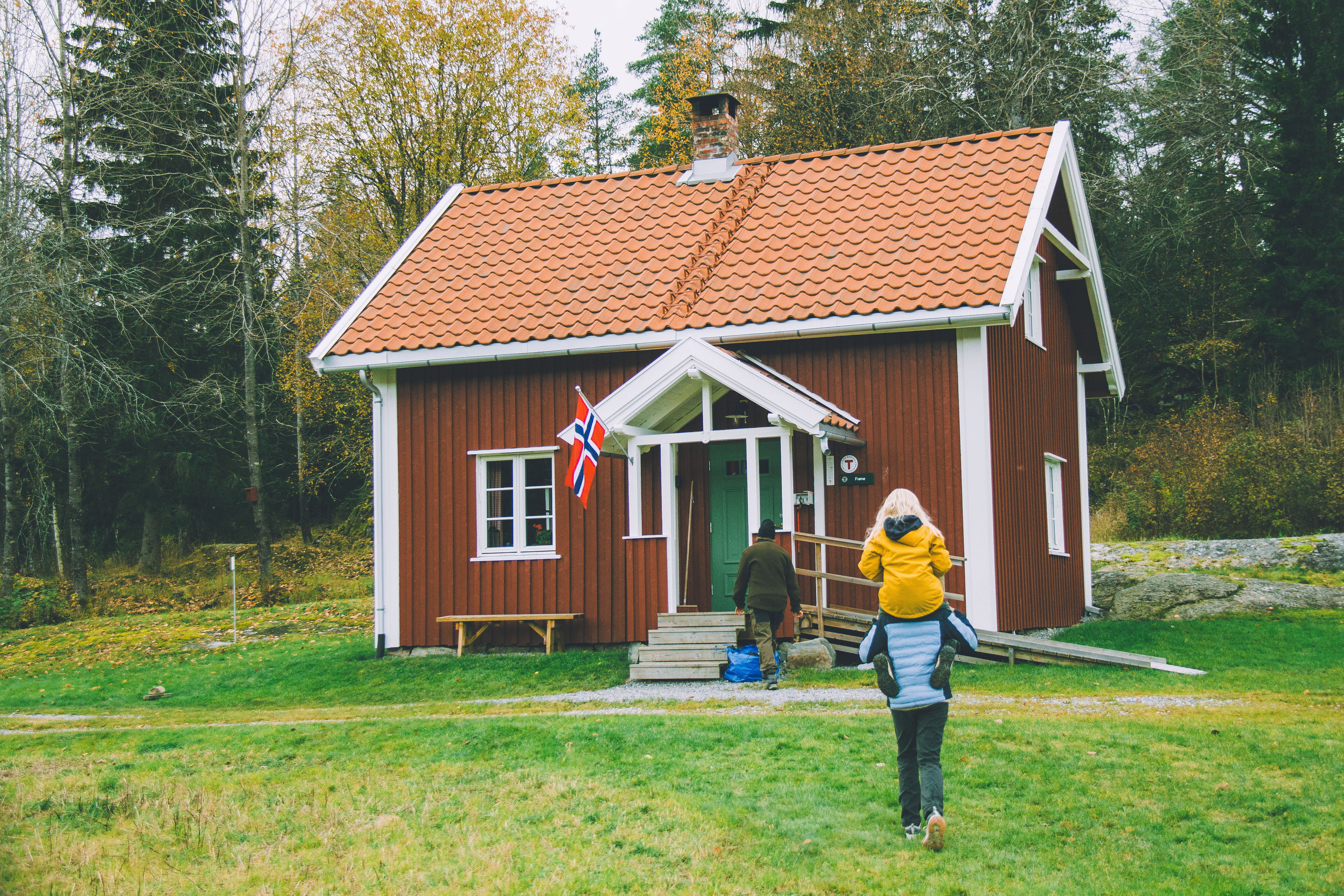 En voksen og to barn g&aring;r mot idyllisk liten r&oslash;dmalt hytte i skogen.