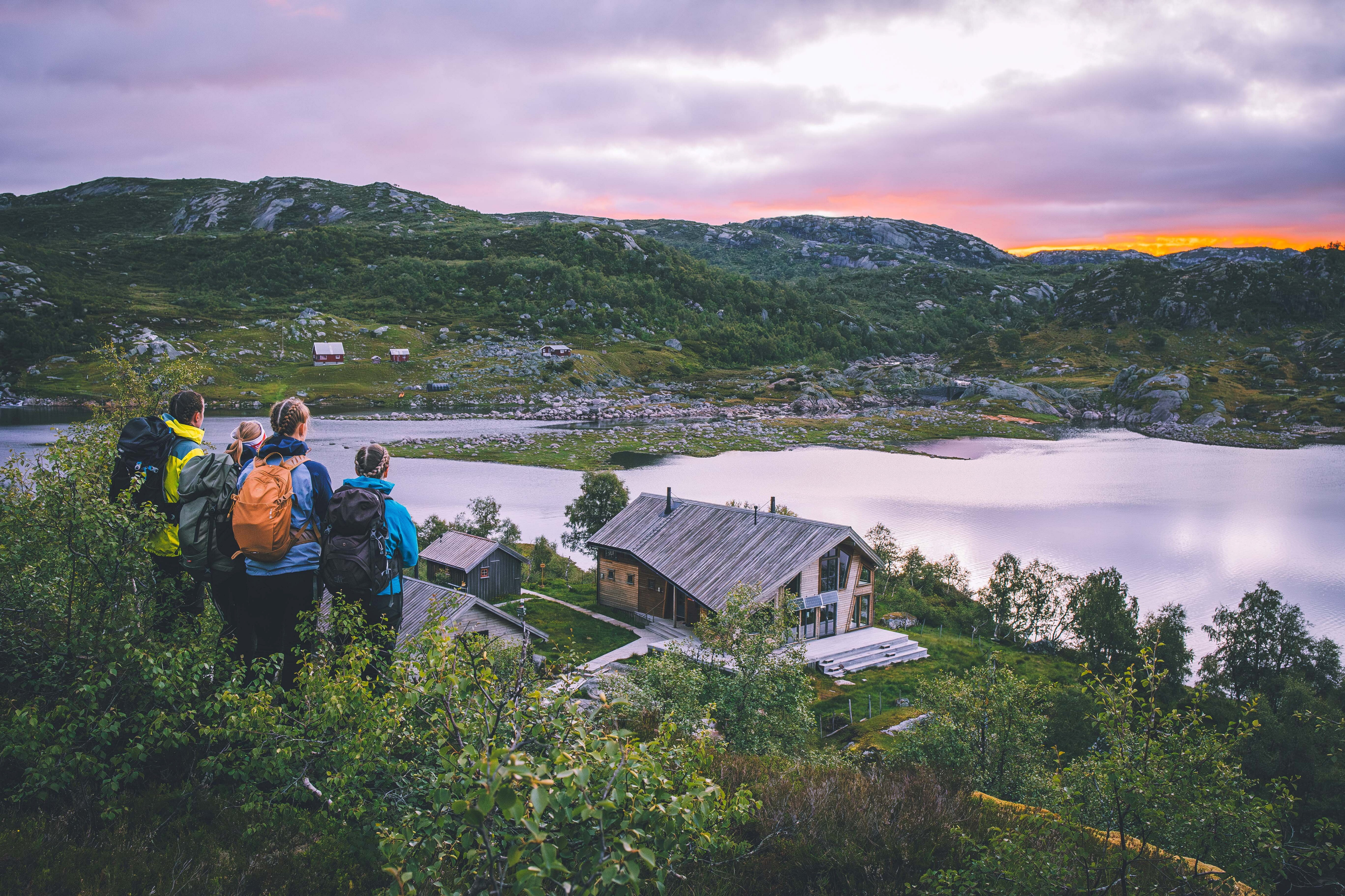 Fire mennesker st&aring;r og ser utover fjellandskap med et vann og en turisthytte.