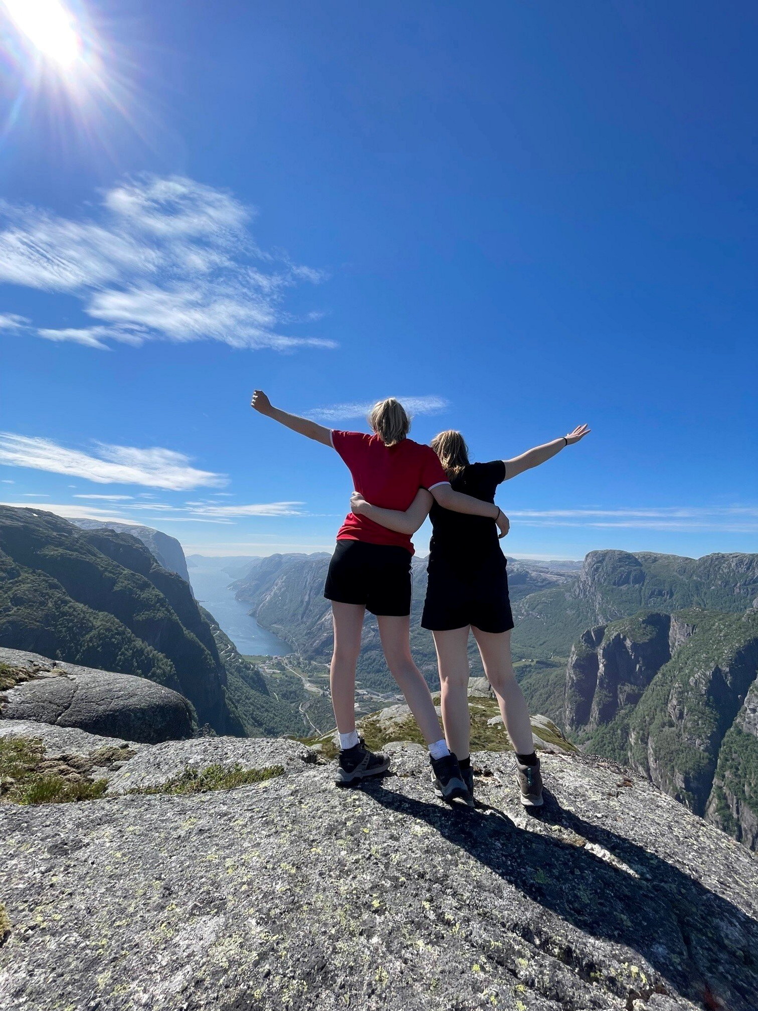 To unge kvinner st&aring;r p&aring; toppen av et fjell og ser utover en fjord.