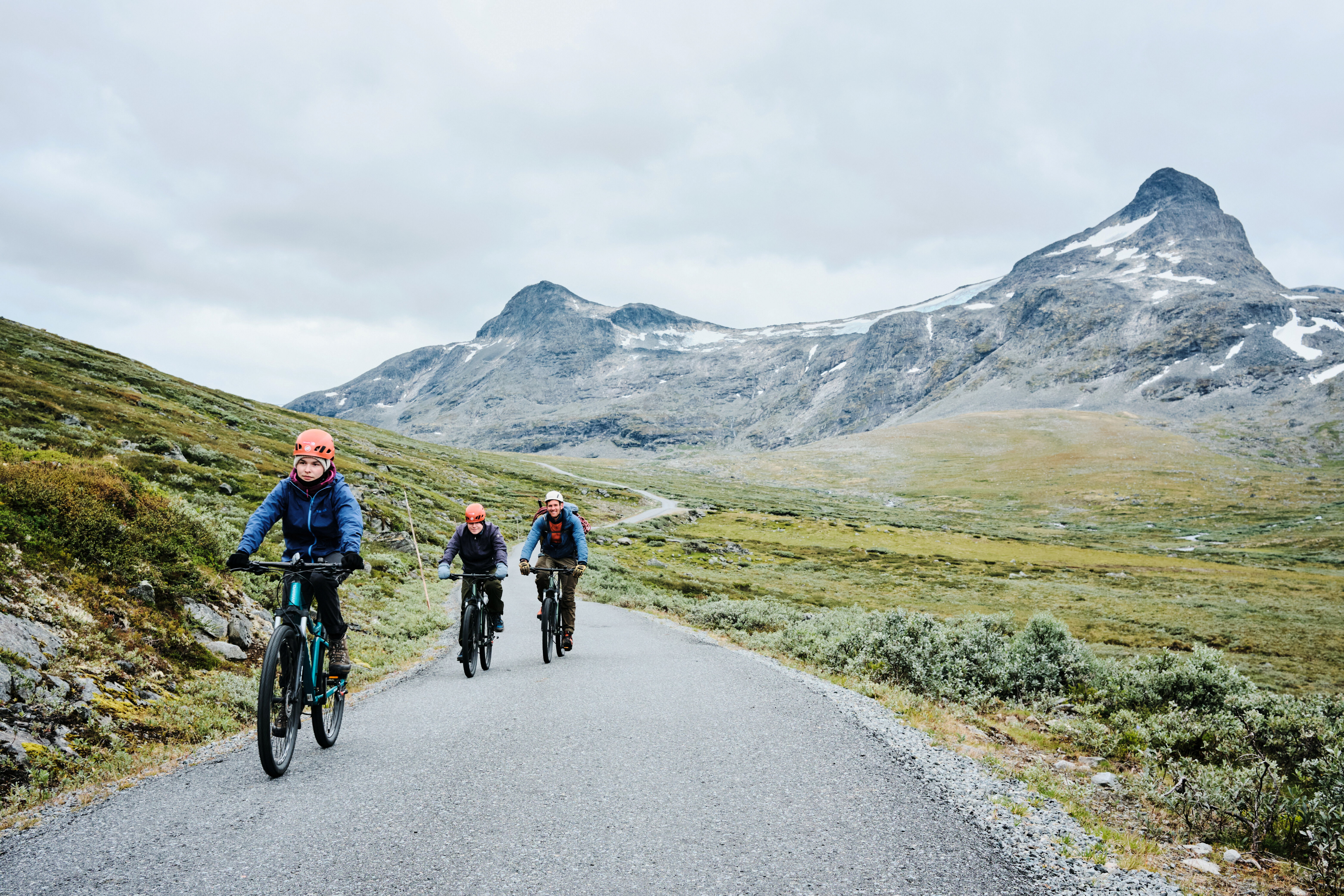 tre personer som sykler p&aring; en vei med fjellandskap rundt seg