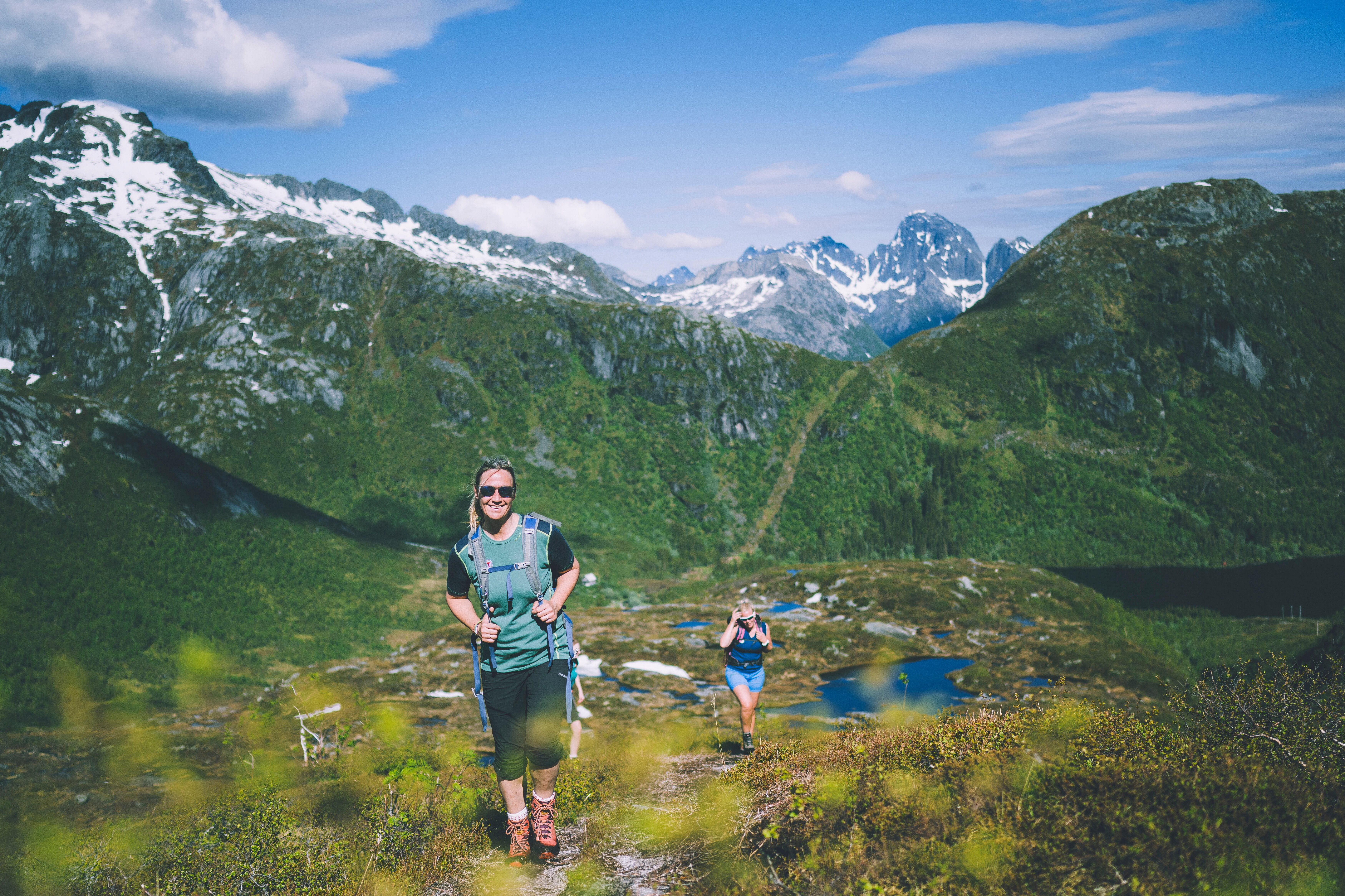 To personer som g&aring;r oppover en sti og smiler mot kameraet. Det er fjell i bakgrunnen