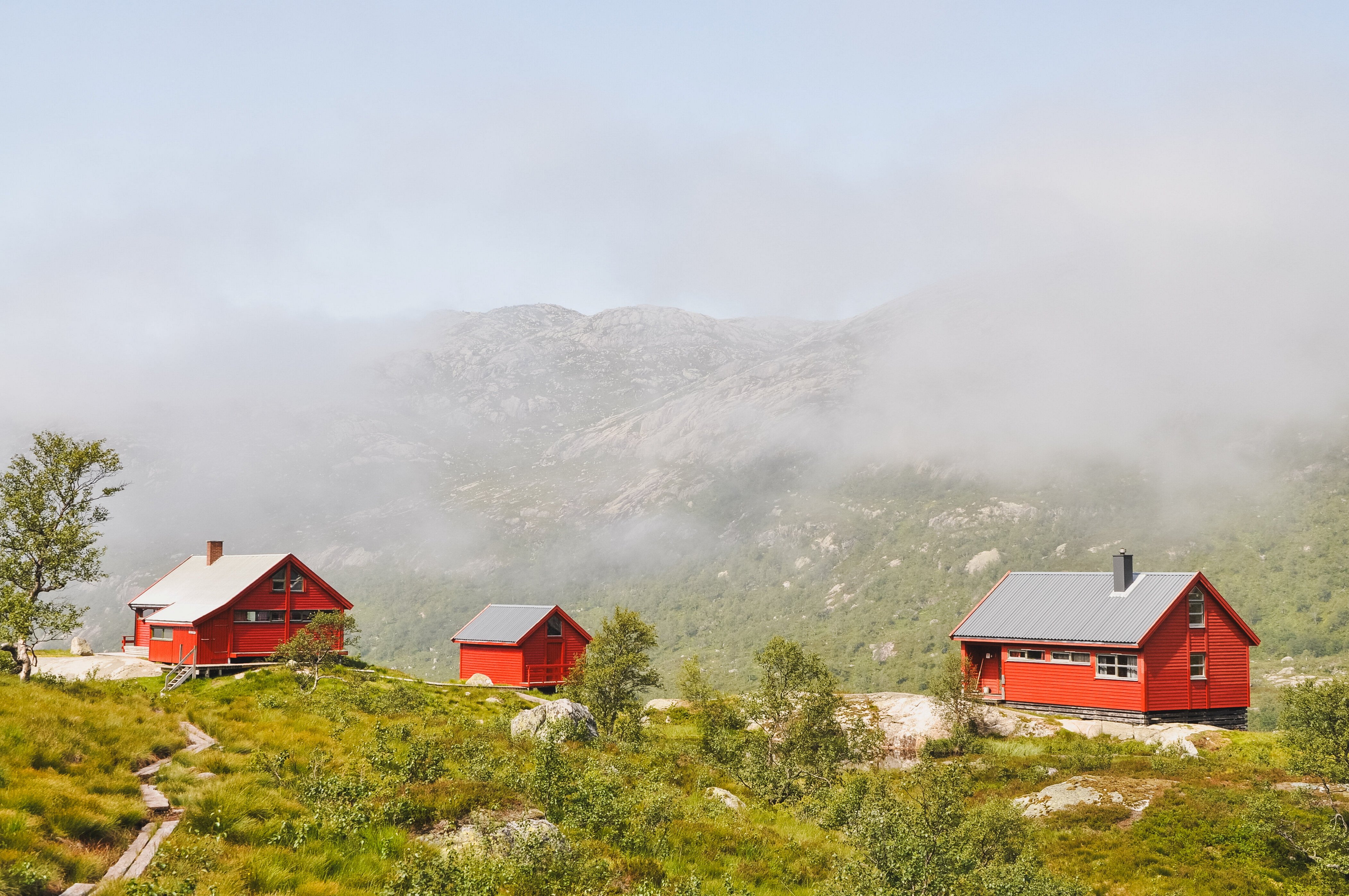 Tre r&oslash;de hytter som er omgitt av t&aring;ke og fjelllandskap.