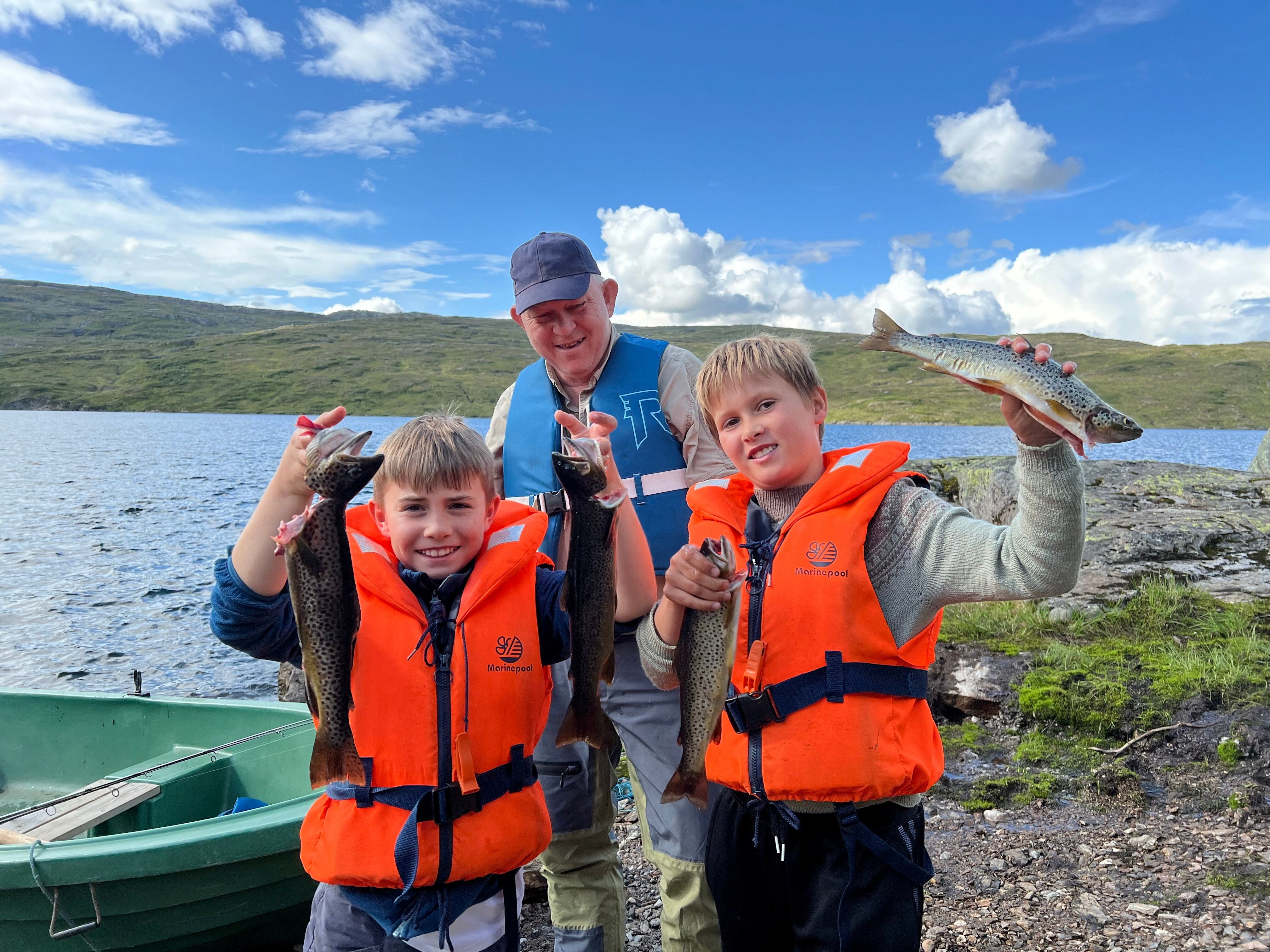 To barn og en mann st&aring;r ved kano ved fjellvann med fisk i hendene